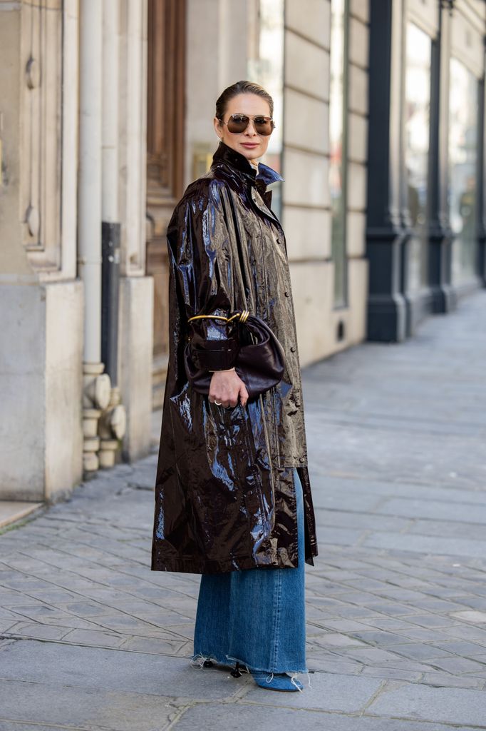 A fashion week guest wears a black vinyl trench coat with jeans and a black bag and sunglasses