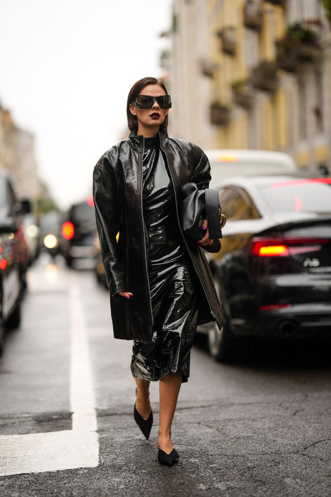 A guest wears black sunglasses, a black shiny varnished leather turtleneck / midi long dress, a black shiny leather oversized coat, black fabric pointed mules from Balenciaga , outside Dolce & Gabbana, during the Milan Fashion Week 