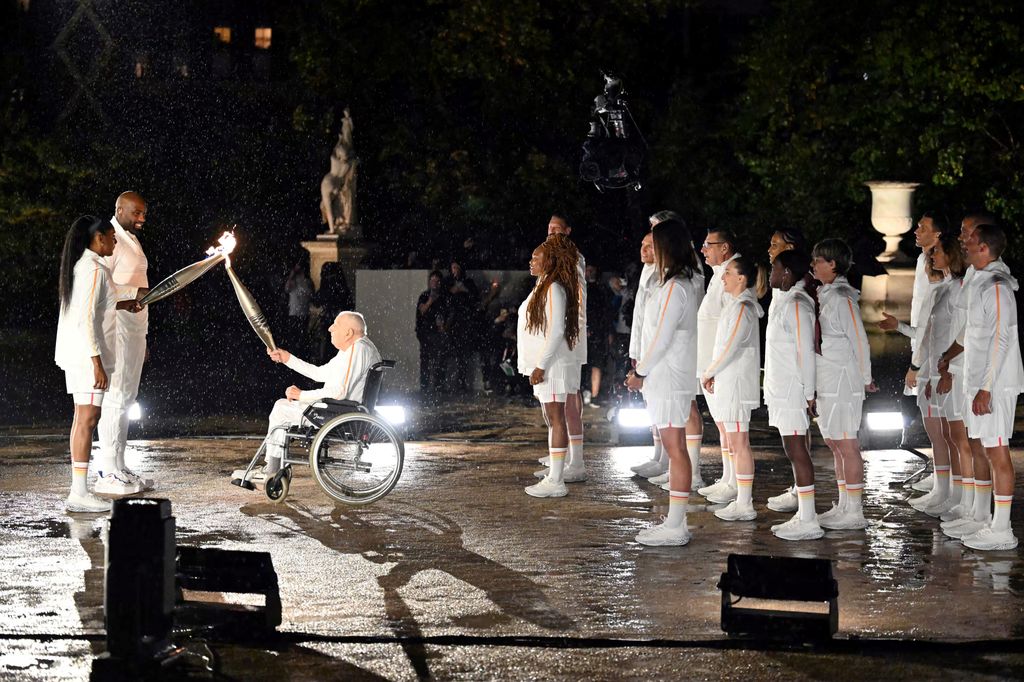 Der 100-jährige Fackelträger und ehemalige französische Radrennfahrer Charles Coste übergibt die olympische Flamme an die ehemalige französische Sprinterin Marie-Jose Perec (li.) und den französischen Judoka Teddy Riner (2. l.).