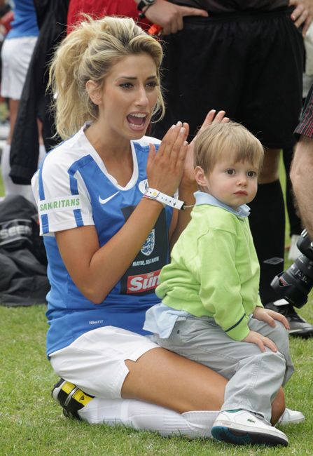 stacey solomon kneels on grass wearing a football shirt as little zachary sits on her lap looking engaged and puzzled by something he is seeing off camera