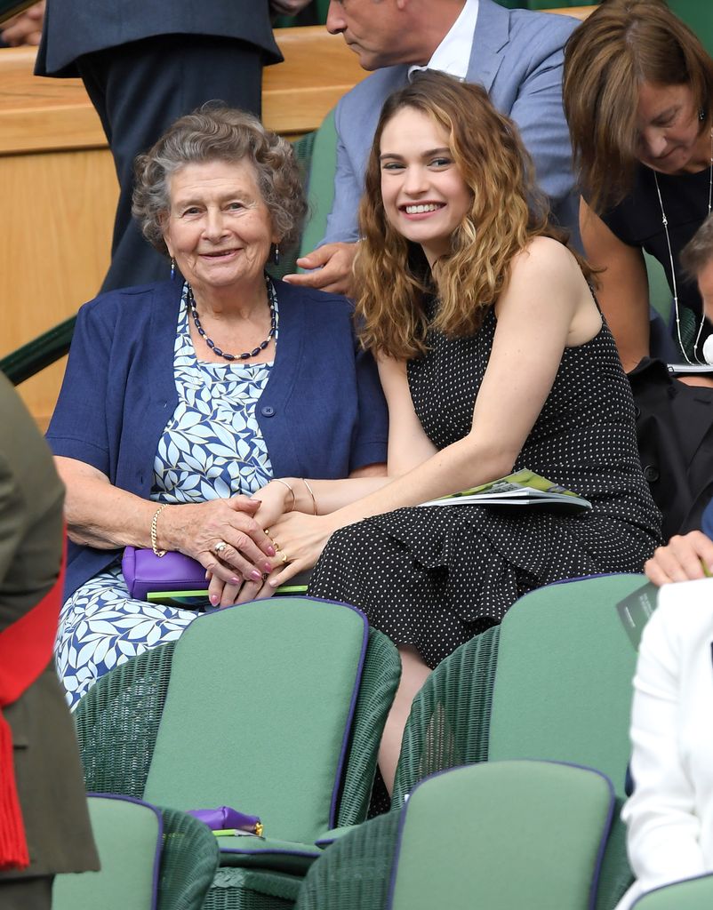 Lily James shares a sweet moment with her grandmother in the Royal Box at Wimbledon 2016. They hold hands, smiling warmly, as they watch the match from green courtside seats—bonding over tennis and timeless elegance.