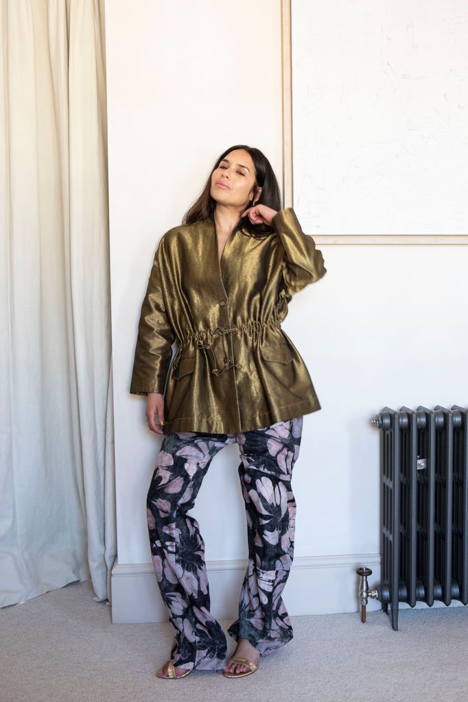 Nieve Tierney stands against a cream wall in a gold metallic drawstring jacket and printed floral trousers, exuding confidence with a soft, closed-eye expression.