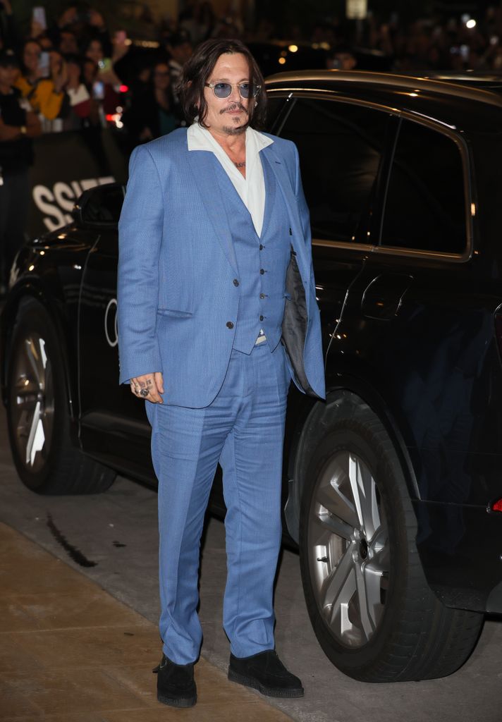 Johnny Depp in a light blue three-piece suit with a white open-collared shirt. They wear round sunglasses, have long dark hair, and are photographed near a black vehicle at night.