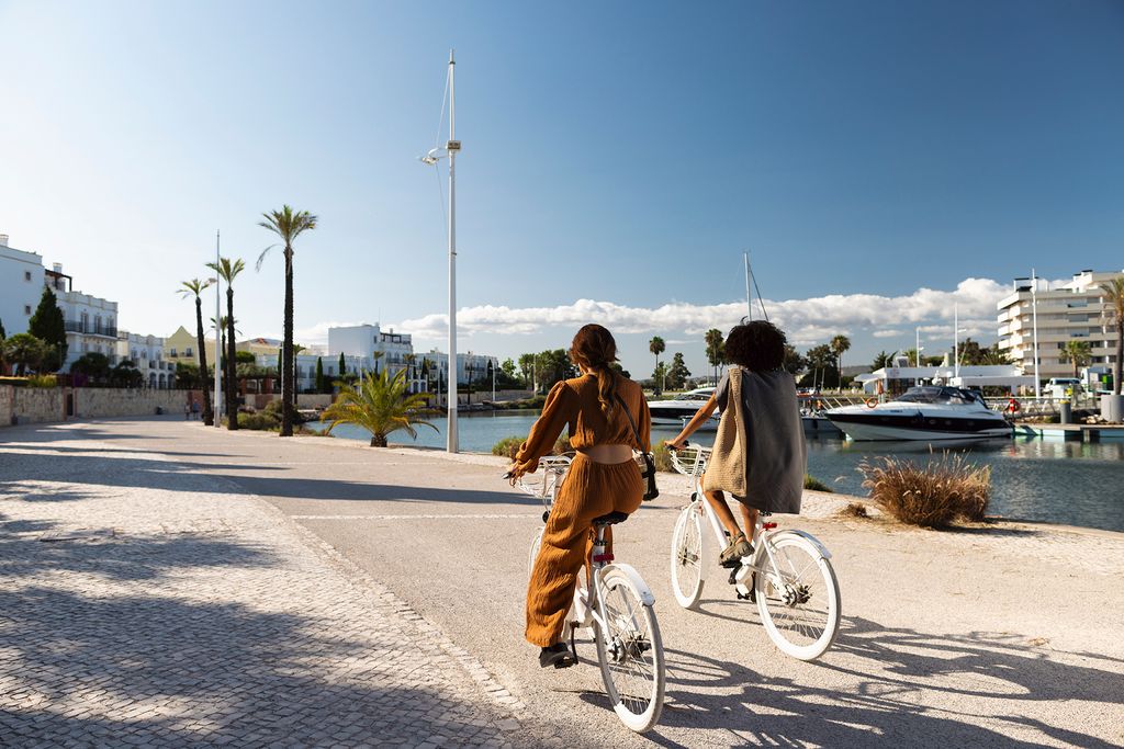 Two women cycling along the waterfront promenade at Vilamoura Marina in Portugal, with yachts docked nearby and modern buildings in the background under a clear blue sky.