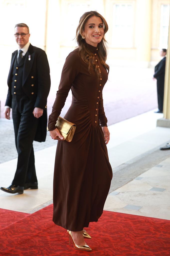 Queen Rania wearing a high-neck brown dress featuring gold buttons, which she paired with a gold clutch and gold heels.