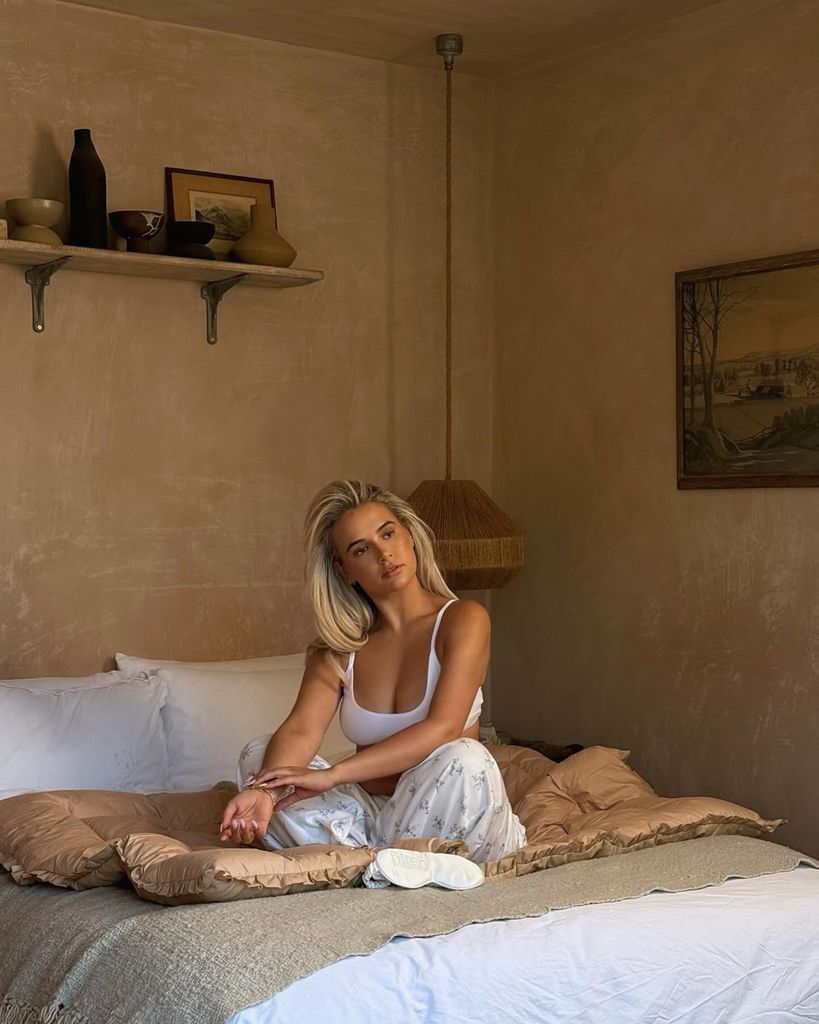 Molly-Mae sits on a hotel bed, she is wearing floral pyjama bottoms and a white bralet. Her hair is blown out and her skin is radiant and glowy.