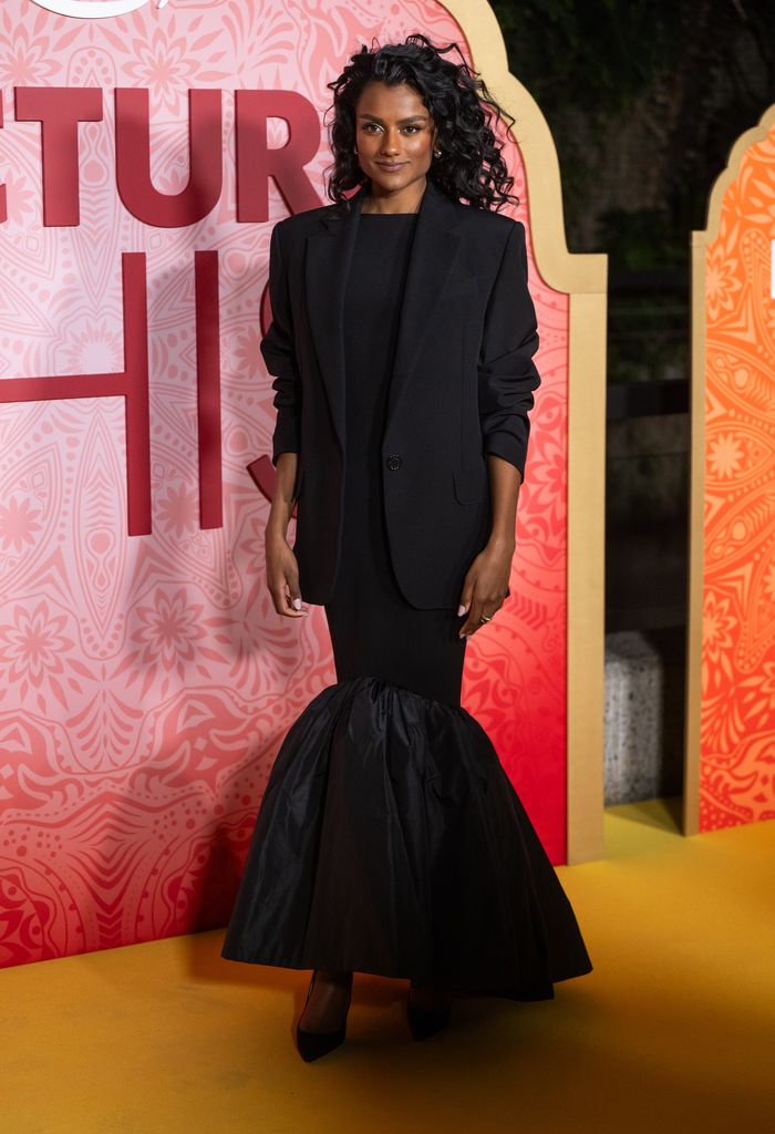 In the image, Simone Ashley stands poised at the premiere of Picture This, against a backdrop adorned with warm, intricate patterns in red, gold, and orange. She wears a sleek, black gown that is fitted at the top before dramatically flaring out at the hem in a voluminous, ruched detail. Over the gown, she dons a sharp, oversized black blazer, adding a structured contrast to the fluidity of her dress. Her hair is styled in natural, voluminous curls that frame her face, and her makeup is soft and glowing with warm tones. She completes the look with classic black heels. Her posture is confident, her gaze steady, and her presence unmistakably commanding—an embodiment of modern elegance and power.