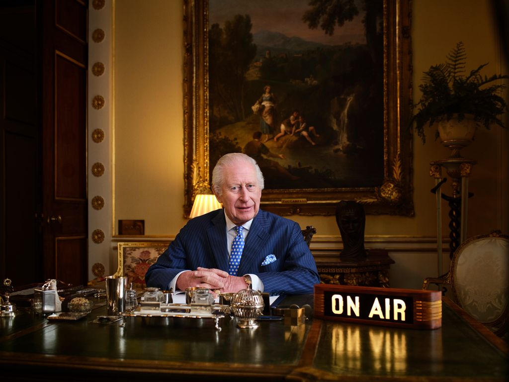 King Charles seated at desk at Buckingham Palace with On Air sign