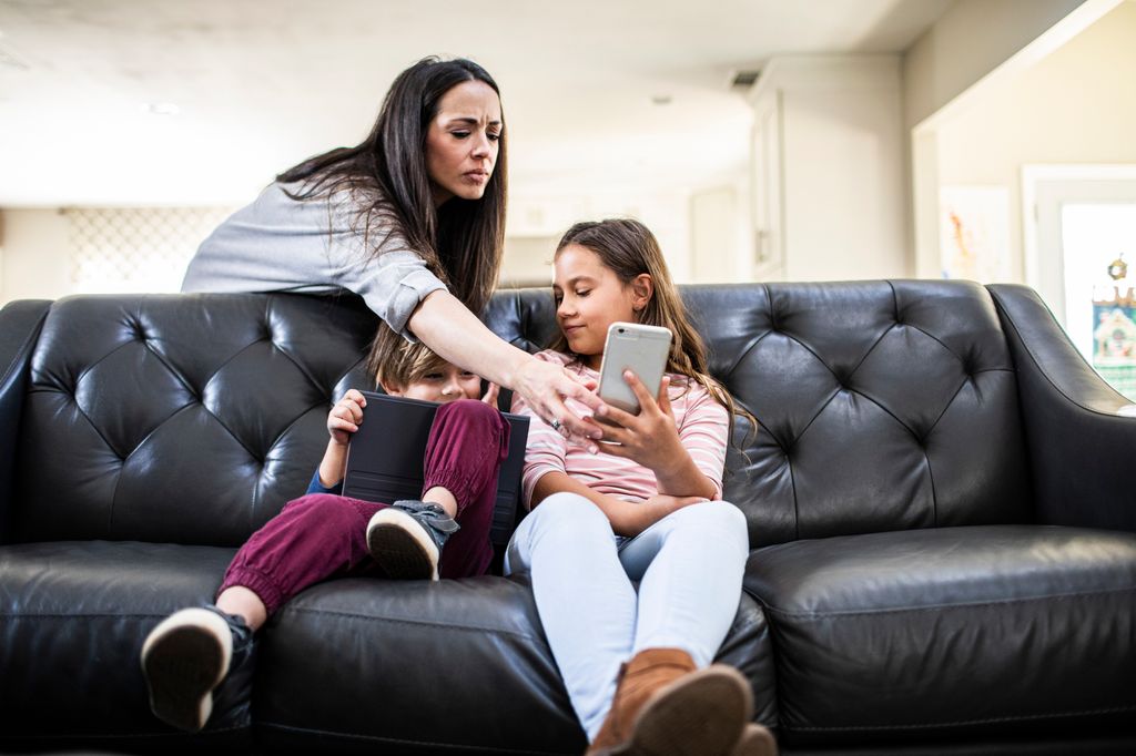 Mother monitoring children using smart devices on sofa at home