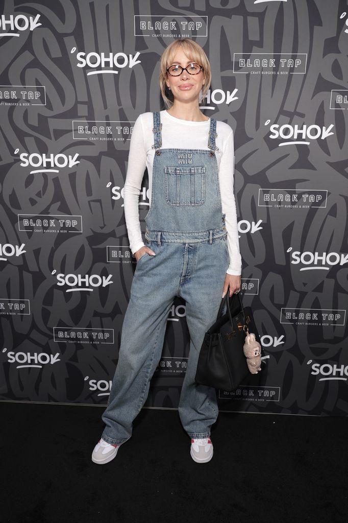 Olivia Attwood wears denim dungarees, white long sleeve tshirt, white trainers and carries a black Hermes bag with bag charm for the event. She wears her hair up and black glasses.