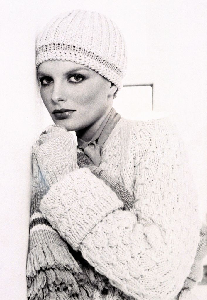 René Russo en noir et blanc portant un bonnet en tricot, un cardigan et des gants reposant sa tête et un bras croisé contre son corps sur un mur.