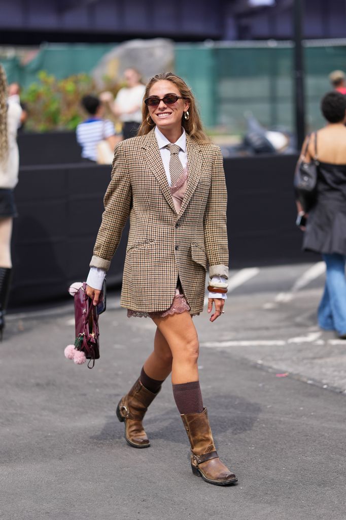 A guest wears long loose light brown hair parted at center, small oval black sunglasses, gold hoop earrings, a small burgundy leather top-handle bag with a pink pom-pom charm, a long single-breasted brown and beige houndstooth blazer jacket with rolled cuffs over a white collared shirt and a brown patterned tie and a dusty pink lace-trimmed mini dress peeking beneath the blazer jacket, dark brown ribbed knee socks and brown leather harness boots with a low stacked heel, outside Coach, during New York Fashion Week, on September 15, 2025 in New York