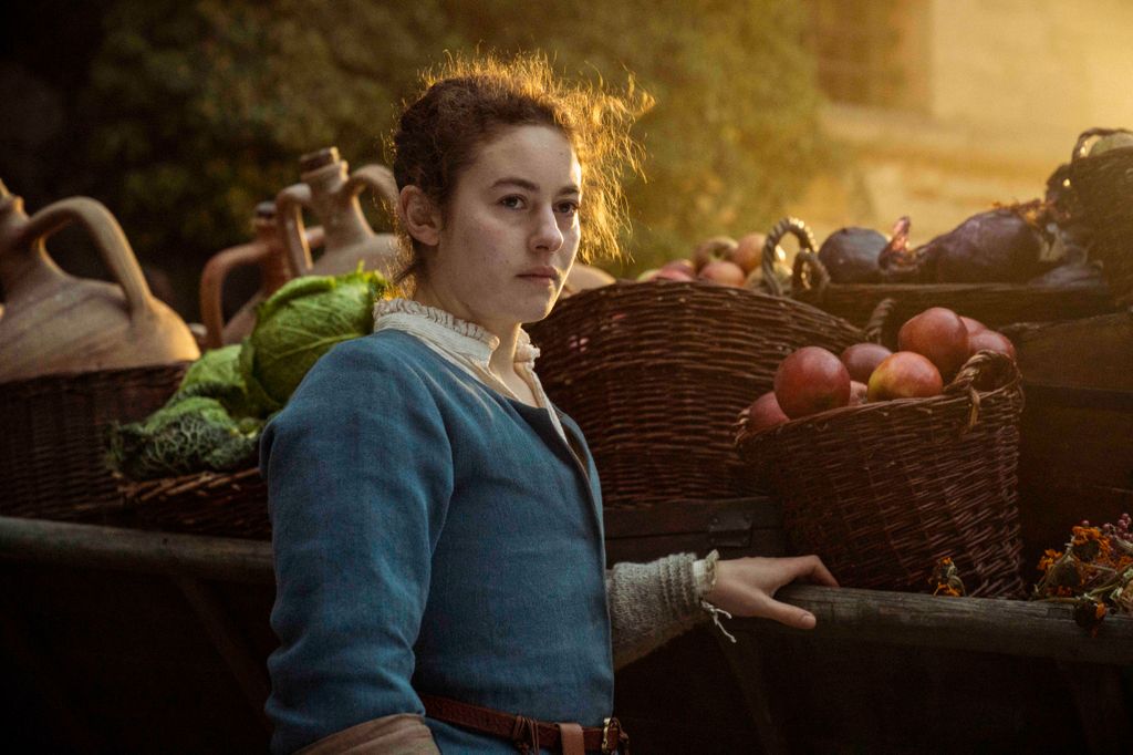 young woman in period dress standing next to baskets of fruit