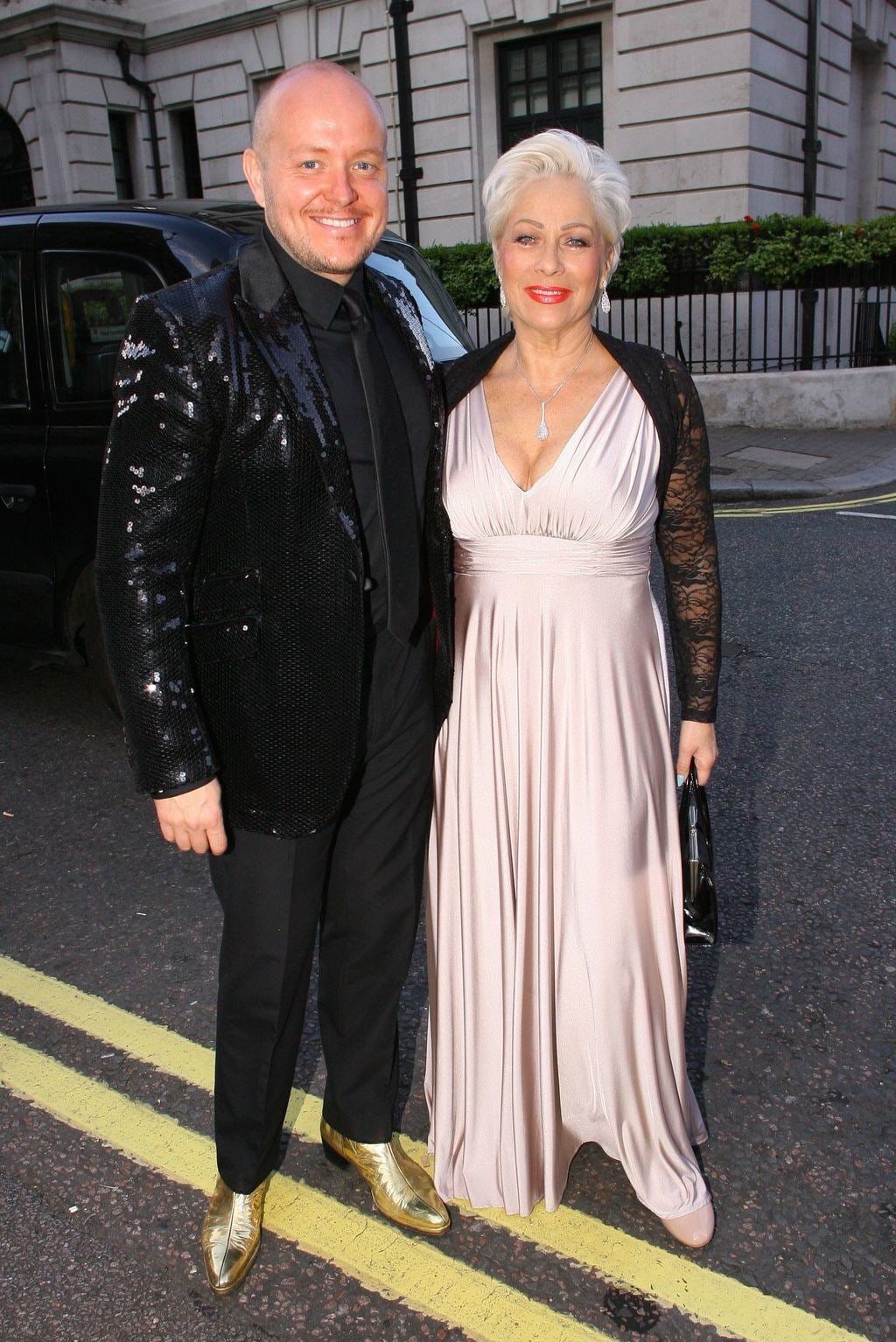 Denise Welch in a satin cream dress with Lincoln Townley in a sequin blazer in the street
