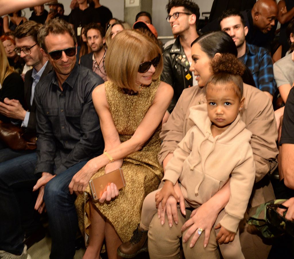 North West sat on Kim's lap at a fashion show, Anna Wintour is chatting to Kim as North looks at the camera