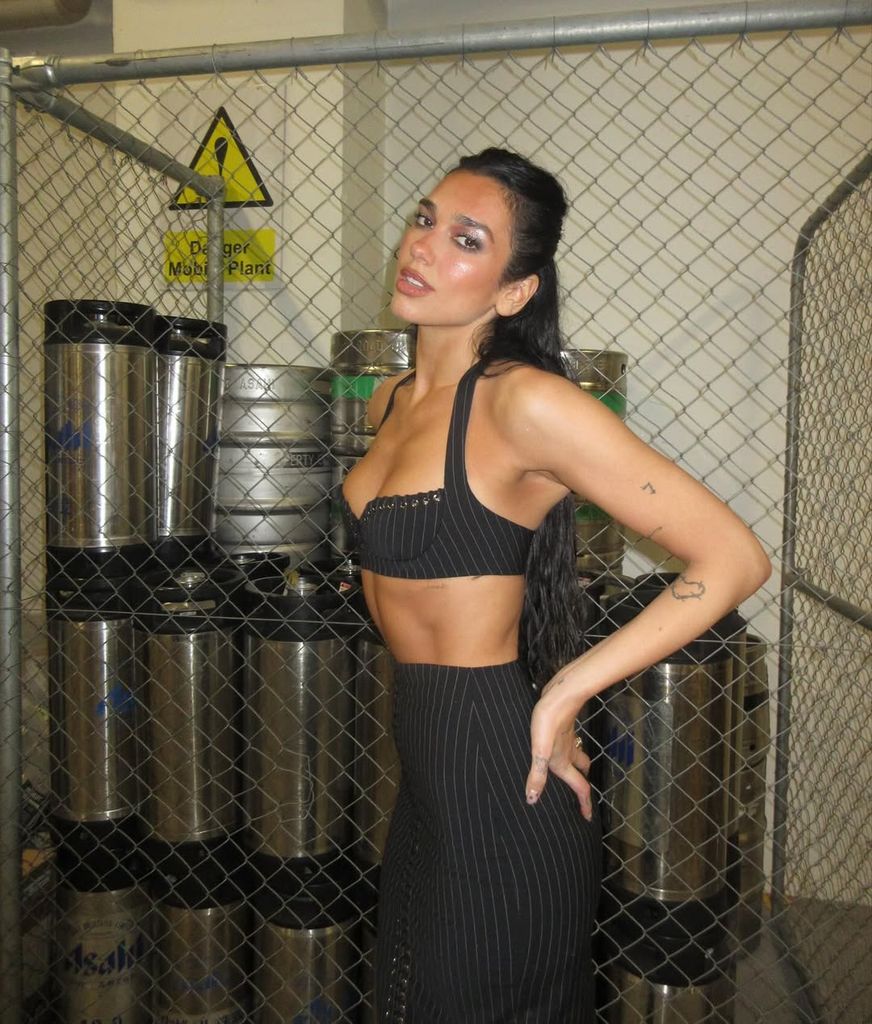 Dua Lipa poses confidently in a side profile shot, wearing a revealing pinstriped co-ord set featuring a halterneck bra and high-waisted lace-up skirt, photographed in an industrial space with metal kegs and chain-link fencing in the background.
