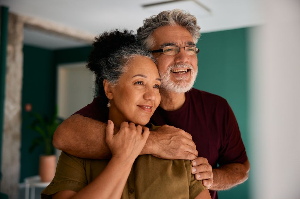 Smiling senior couple embracing each other while gazing away with hope and love, savoring a peaceful moment together in their cozy home filled with warmth and comfort