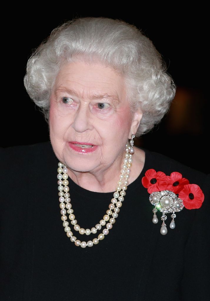 Queen Elizabeth II wears a three-strand pearl necklace with a poppy brooch. She looks serious yet elegant, with her iconic white hair and regal demeanor.