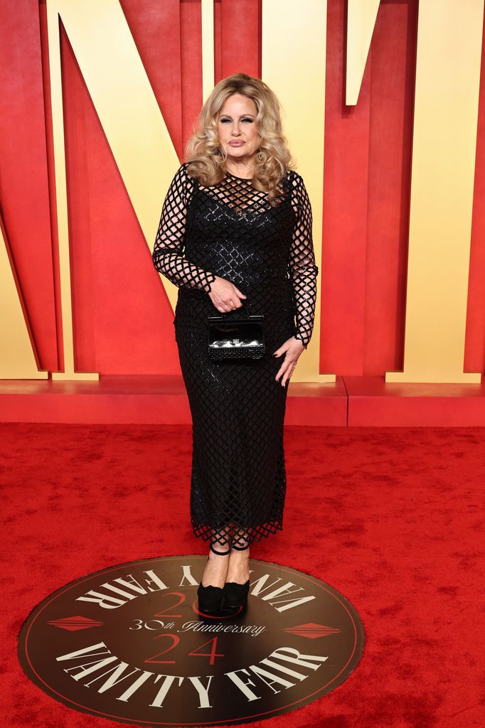 Jennifer Coolidge wears a black long sleeve maxi dress in black sequin fishnet, with black closed-toe heels and a black top-handle handbag.