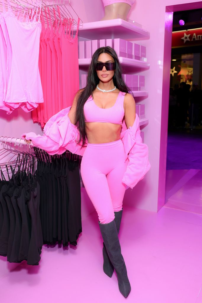 Kim Kardashian poses near some of her shapewear, she is wearing sunglasses and is dressed in bright pink