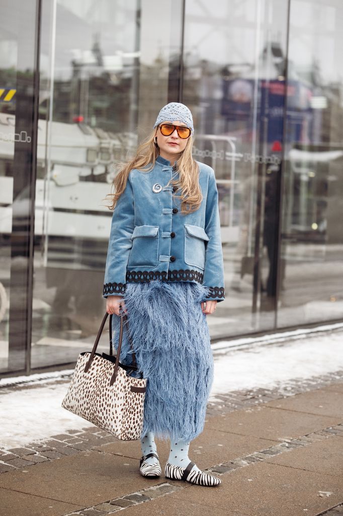 A guest wears blue fur skirt, blue jacket, blue socks with polka dots, animal print bag and zebra print shoes, light blue knitted hat and orange sunglasses outside the OperaSport show