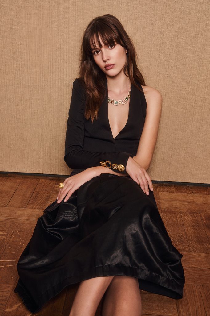 Ruby Sear sits elegantly in a black asymmetric Schiaparelli dress, paired with a shimmering Bucherer necklace, gold buttons, and a statement ring. The setting exudes understated luxury.