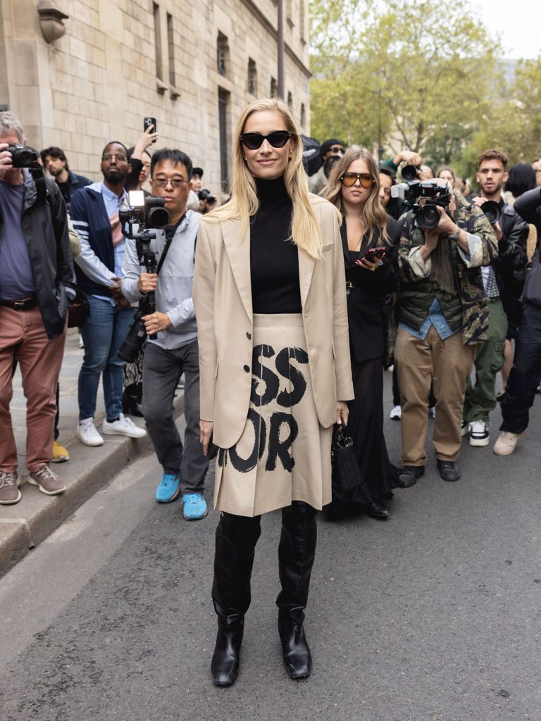 Beatrice Borromeo poses in a tan trench with bold black Dior lettering, styled with a turtleneck, boots and cat-eye sunglasses outside Paris Fashion Week.