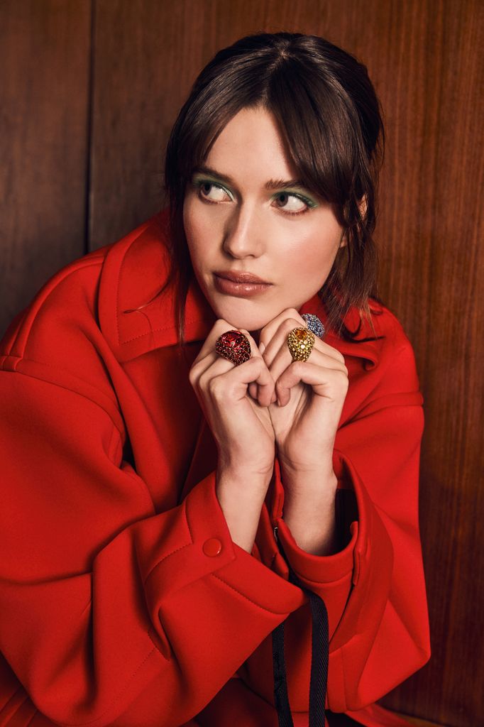 Ruby Sear dons a bold red coat from Toga with dramatic green eyeshadow, accessorized with oversized, vibrant Bucherer rings. Her pose is contemplative, emphasizing the striking jewelry.