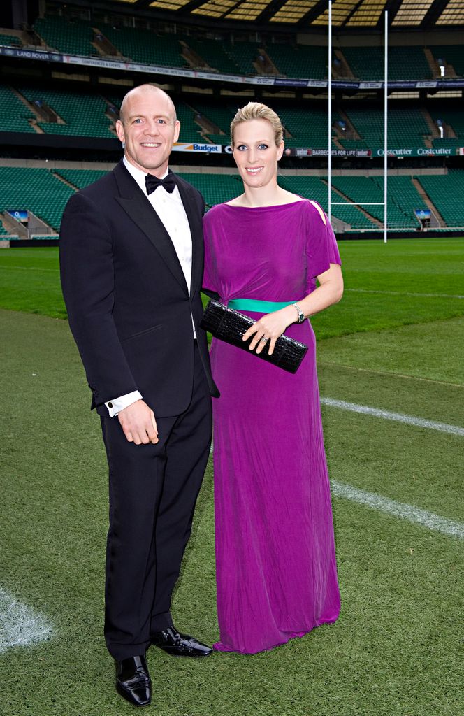 Mike Tindall in a suit and Zara Tindall in a purple dress. The pair are stood on a rugby pitch