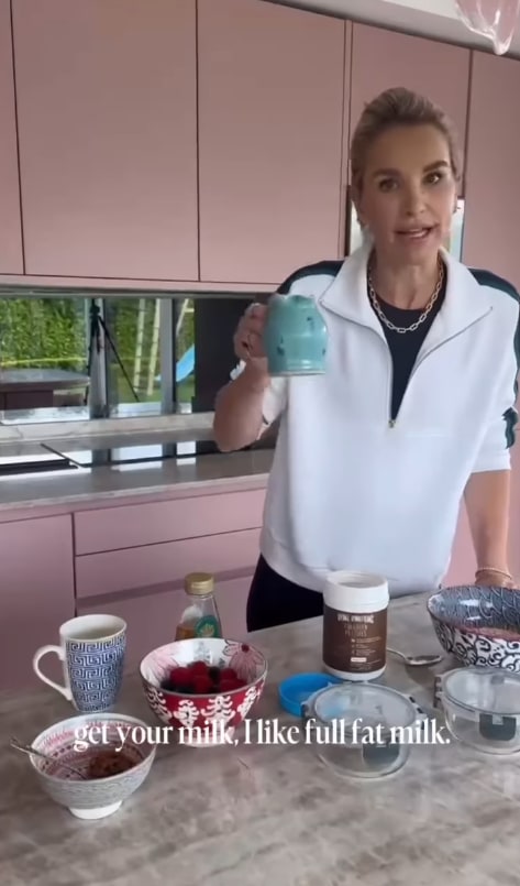 A woman with blonde hair in a ponytail, wearing a white jacket and chain necklace, holds up a light blue mug in a modern kitchen with pink cabinets.