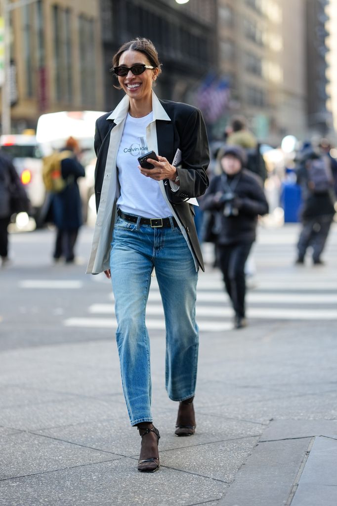 A guest wears black sunglasses, white Calvin Klein Jeans print t-shirt, cream unbuttoned long sleeve shirt, black blazer jacket, black leather belt, navy blue washed straight denim jean pants, black sheer mesh tights, black heels leather shoes, outside Calvin Klein, during the New York Fashion week Fall/Winter 2025 on February 7, 2025 in New York, New York. (Photo by Edward Berthelot/Getty Images)