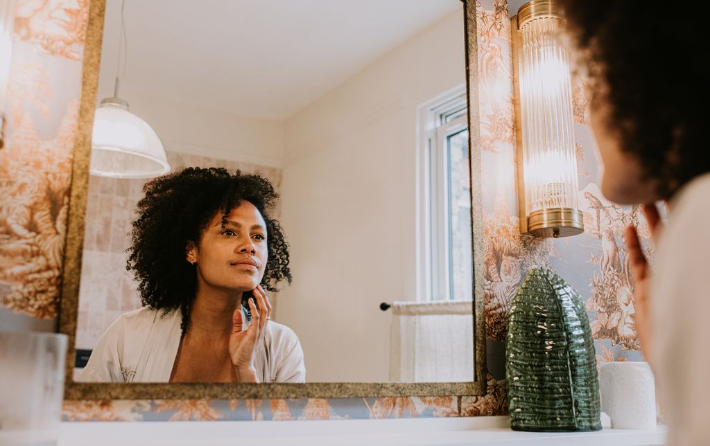 A beautiful black woman looks into a bathroom mirror and gently touches her skin as she examines her reflection.