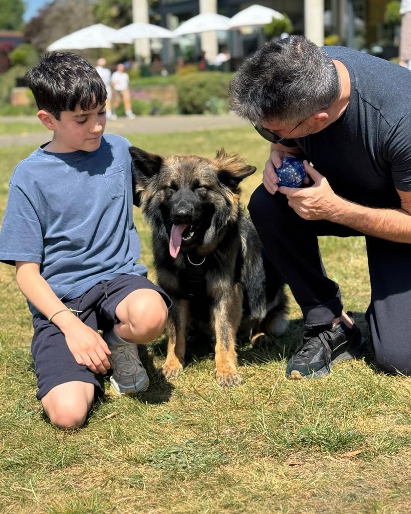Eric and Simon Cowell kneeling beside their German shepherd dog after a walk in a public park