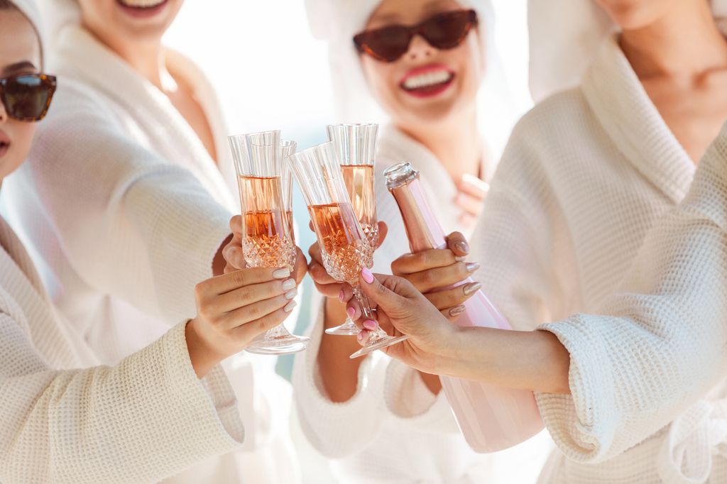 female friends in white bathrobes before a bachelorette party toasting with champagne