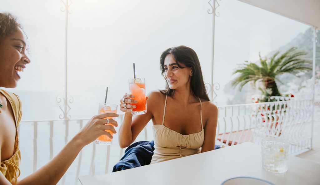 Two females on holiday, enjoy a cool, refreshing drink. They clink their glasses together before they take a sip. Image depicts dining, restaurants, vacationing, experiences and alcoholic and non-alcoholic drinks.