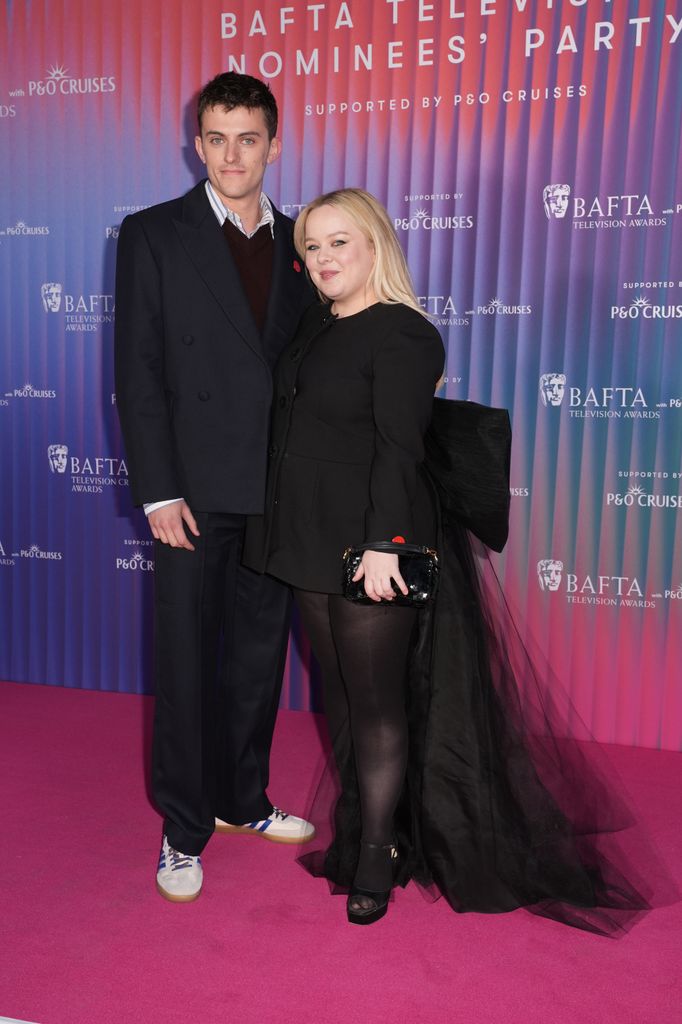 Jake Dunn stands next to Nicola Coughlan on the pink carpet. He wears a blazer, teamed with a jumper and shirt, black trousers and trainers. 