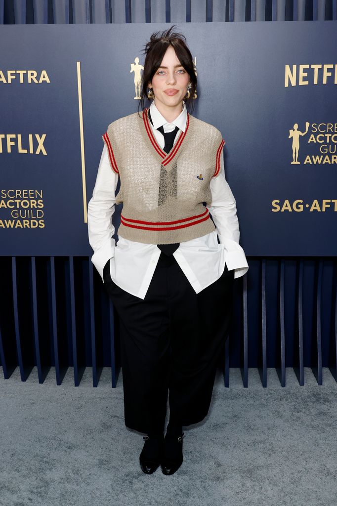 Billie dons a preppy Vivienne Westwood outfit with a beige knit vest, oversized white shirt, and wide black trousers. Her hair is casually styled, adding effortless cool to her look.