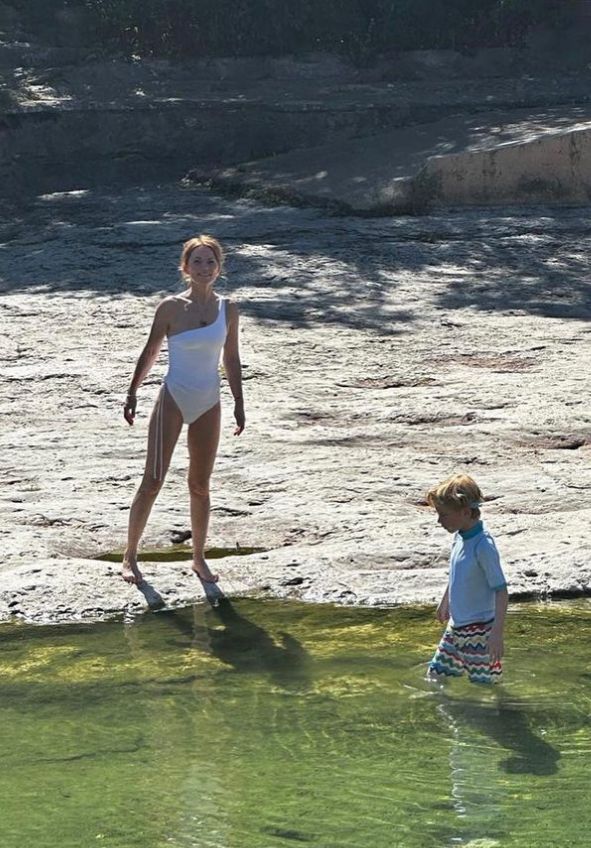 Geri Halliwell in a white swimsuit on edge of water with a young boy in blue shirt and shorts in the water