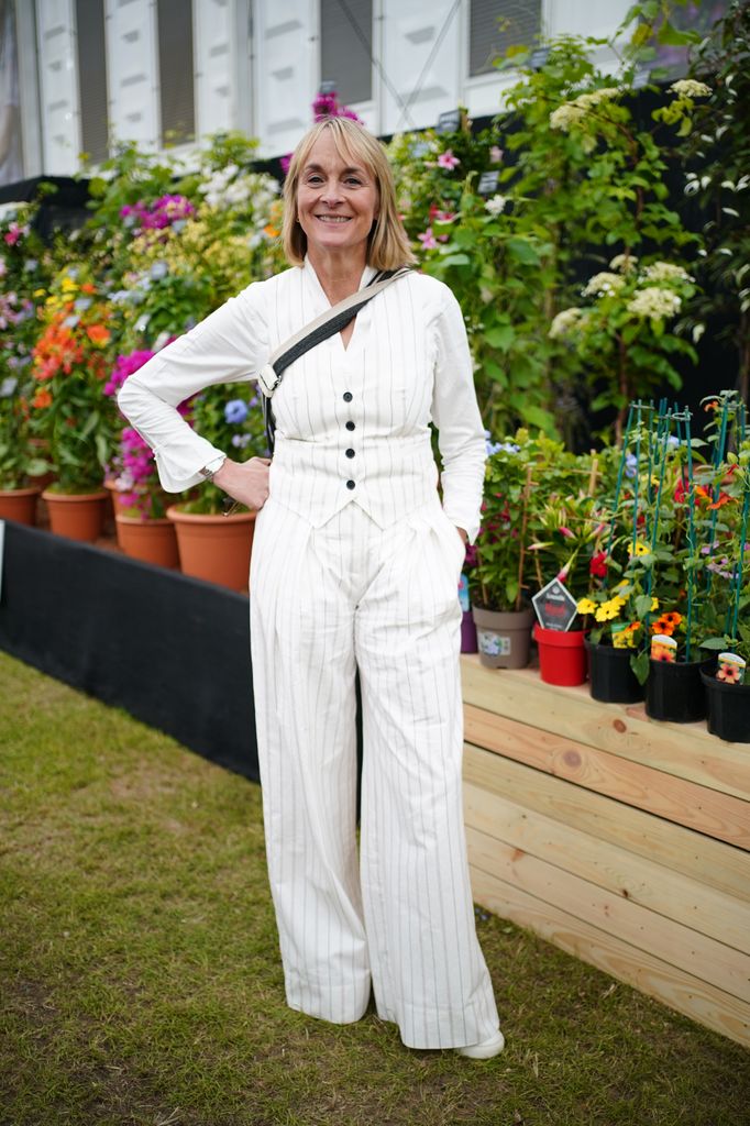 Louise Minchin in a white pinstripe waistcoat and wide-leg trousers smiles in front of a flower display at Chelsea Flower Show 2025.