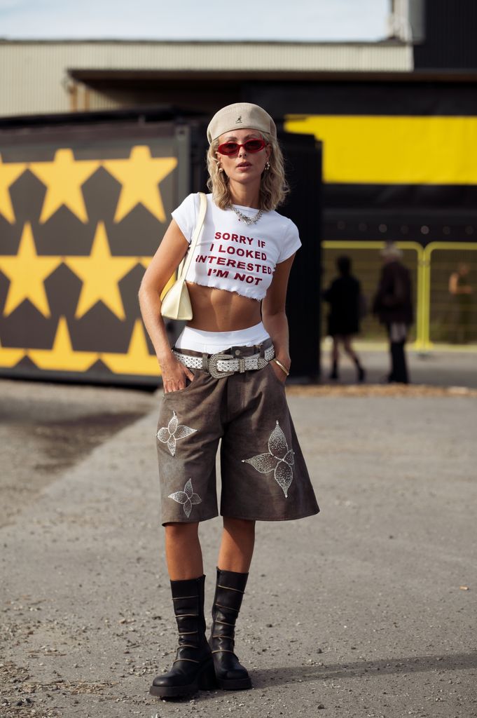  A guest wears brown wide shorts with white belt, white cropped shirts with printed text in red letters, white underwear, beige hat and black long boots outside the Rolf Ekroth s show