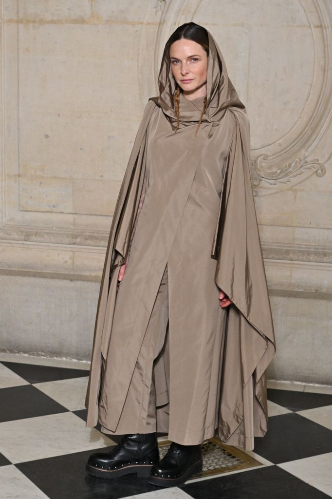 Rebecca rocks a hooded taupe cape and platform boots, bringing avant-garde fashion drama to a Dior event.