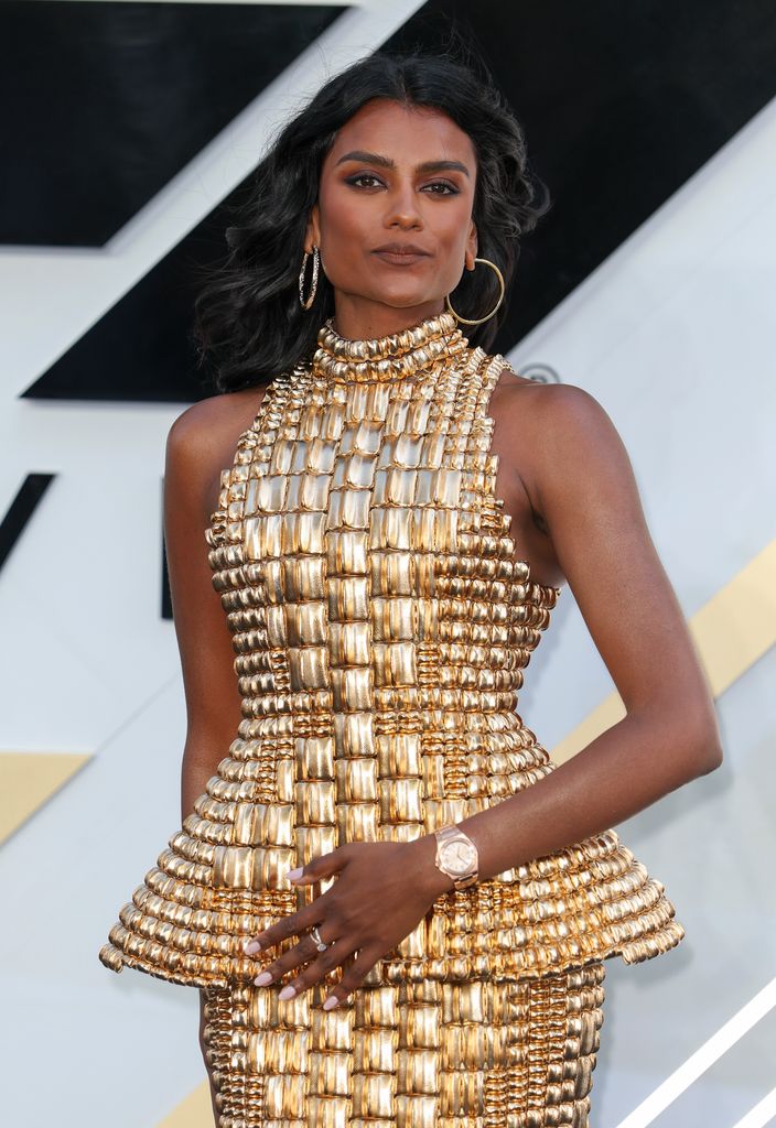 Simone Ashley, a British-Indian actress with warm brown skin and long, softly waved black hair, stands poised and confident against a modern monochrome and gold backdrop. She wears a figure-hugging, futuristic gold dress that appears to be constructed from small, metallic, rectangular tiles—each catching the light like a jewel. The structured dress flares at the waist with a peplum detail, enhancing her hourglass silhouette, and extends into a form-fitting pencil skirt that ends mid-calf. Her neckline is high and halter-style, lending a regal air to the ensemble. She accessorizes with large gold hoop earrings and gold strappy heels that perfectly complement the ensemble’s opulence. Her makeup is minimal but luminous—bronzed skin, soft smoky eyes, and a neutral lip—allowing the dress to command center stage.