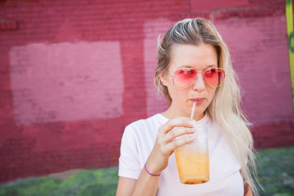 Jeune femme blonde portant des lunettes de soleil roses buvant du jus dans une tasse en plastique et de la paille devant un mur rose