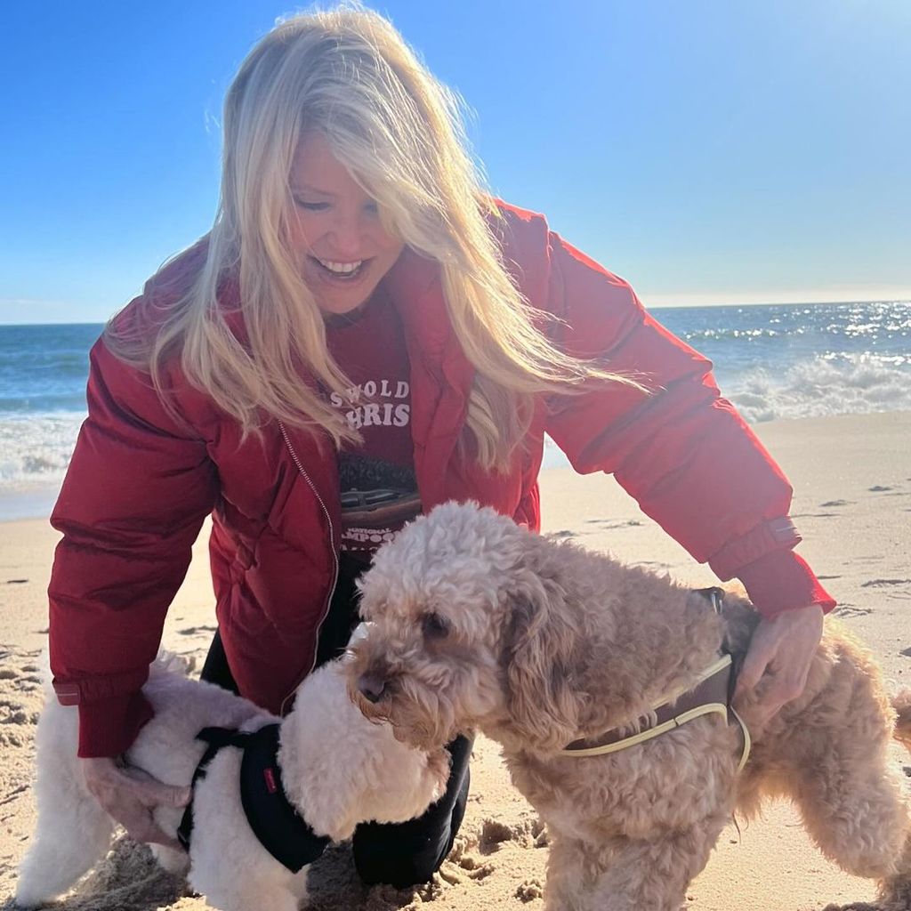 Christie Brinkley kneels in the sand and holds onto her two puppies
