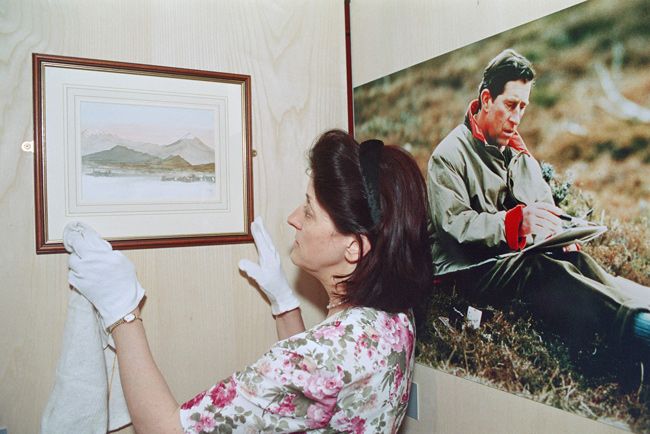 a gallery employee carefully hangs a watercolour painting of a landscape on a wall beside a framed photograph of king charles sitting in long grass holding a brush to a page