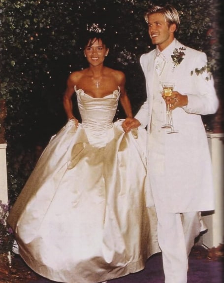 Victoria smiling in her white wedding dress and David next to her in his white suit holding a glass of fizz. 