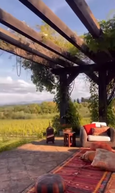 A cozy outdoor seating area under a wooden pergola with vines, featuring a cushioned chair, a small table, a patterned rug, and scenic views of green vineyards and distant hills under a partly cloudy sky.