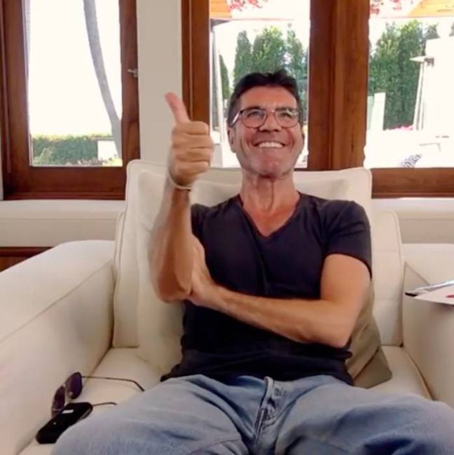 simon sits on a cream sofa in front of a window where cacti grow and his gives a thumbs up signal 