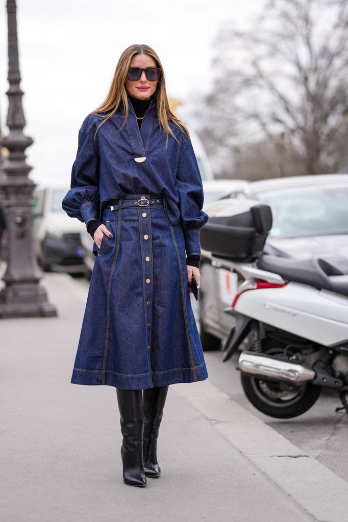 Olivia Palermo wears brown marble print pattern sunglasses, a gold and green large chain necklace, a black ribbed wool turtleneck pullover, a navy blue denim oversized V-neck / puffy sleeves shirt, a matching navy blue denim / gold buttoned / midi long skirt, black shiny leather pointed heels outside Zimmermann, during Paris Fashion Week 