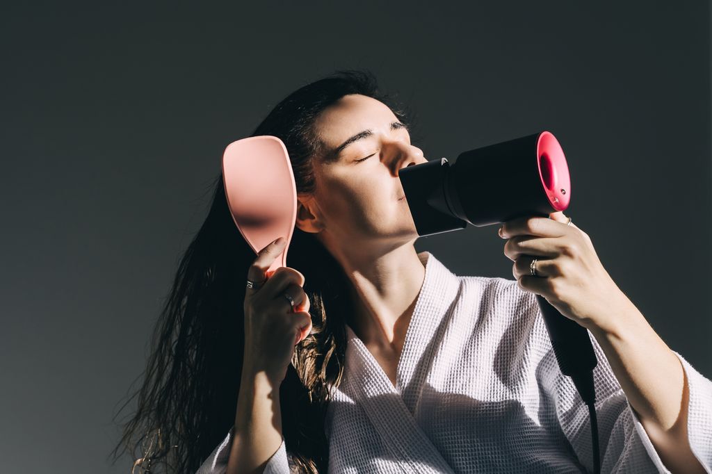 A young woman in white bathrobe blow-dries her hair at home.